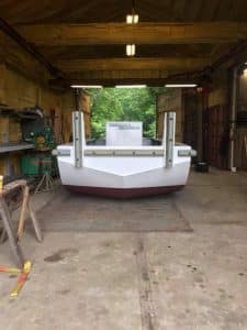 Fitzgerald Metal Works. Truckable sectional barge with white and maroon steel hull in a workshop garage. The barge is positioned near an open doorway leading to green foliage outside.