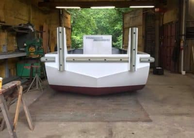 Fitzgerald Metal Works. Truckable sectional barge with white and maroon steel hull in a workshop garage. The barge is positioned near an open doorway leading to green foliage outside.