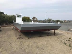 Fitzgerald Metal Works. A newly constructed sectional barge with a steel hull, displayed on supports in a shipyard. White wheelhouse with a gray deck and maroon underneath.