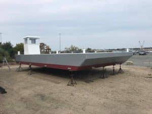 Fitzgerald Metal Works. A newly constructed sectional barge with a steel hull, displayed on supports in a shipyard. White wheelhouse with a gray deck and maroon underneath.