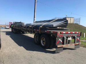 Fitzgerald Metal Works. A flatbed semi-truck loaded with large, black metal panels. The truck is parked on a concrete surface under clear blue skies.