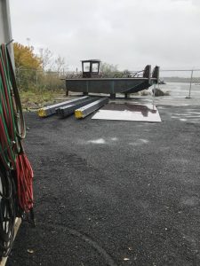 Fitzgerald Metal Works. Custom Boats. large metal barge on land. Metal beams are stacked on the ground beside the barge.