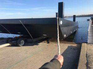 Fitzgerald Metal Works. A large black barge on a trailer at the water's edge, with a hand holding a mooring rope.