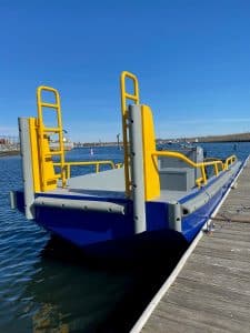 Fitzgerald Metalworks. A blue, yellow and grey small utility barge or workboat is docked at a marina.