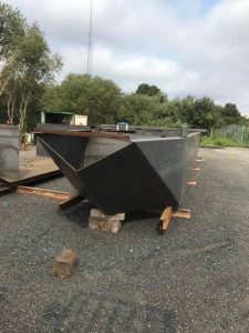 Fitzgerald Metal Works. A large, dark gray Steel Boat Hull with an angular design sits on wooden supports