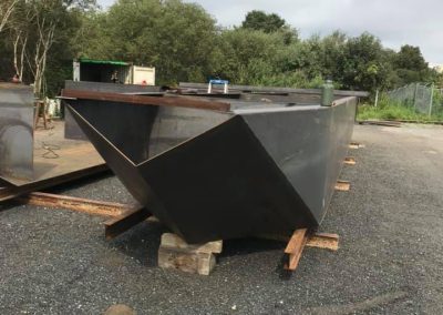 Fitzgerald Metal Works. A large, dark gray Steel Boat Hull with an angular design sits on wooden supports