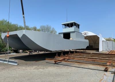 Fitzgerald Metal Works. Large grey barge on stilts in an industrial yard. Steel beam being lifted by a crane onto barge.