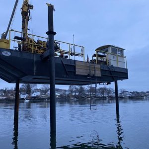 Fitzgerald Metal Works. Large barge being lowered into water. Yellow machinery on barge as well as a ladder going down on the side of boat.