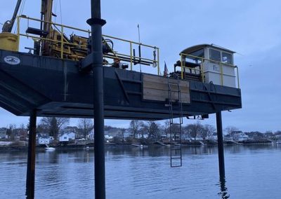 Fitzgerald Metal Works. Large barge being lowered into water. Yellow machinery on barge as well as a ladder going down on the side of boat.