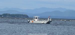 Fitzgerald Metalworks. A small workboat or utility barge on a body of water, with a scenic backdrop of distant mountains and an island covered with trees.