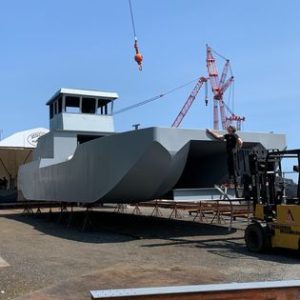 Fitzgerald Metalworks. Large grey barge on stands. Orange crane above barge and worker in a yellow machine at the hull of boat.