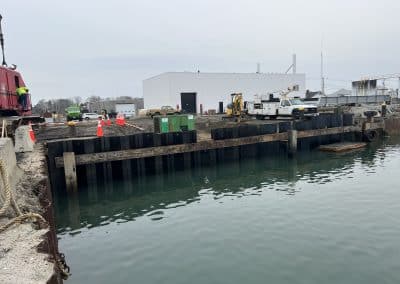 Fitzgerald Metalworks. Marine Construction. Black metal sheets line the waters edge. Buildings, cars and machines located in the background.