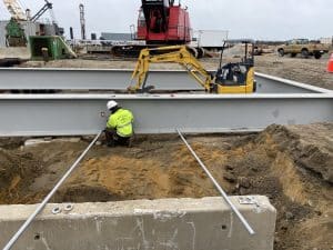 Fitzgerald Metal Works. Large steel beam being placed in the ground. Yellow machine and worker in yellow jacket welding metal.