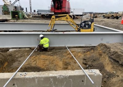 Fitzgerald Metal Works. Large steel beam being placed in the ground. Yellow machine and worker in yellow jacket welding metal.